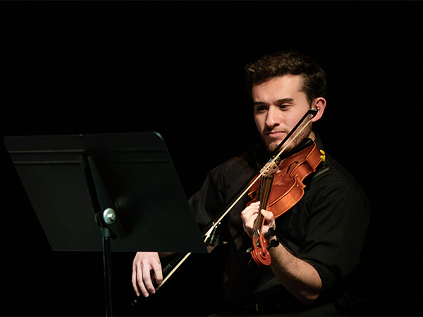 A male student plays violin in a dark performance space.